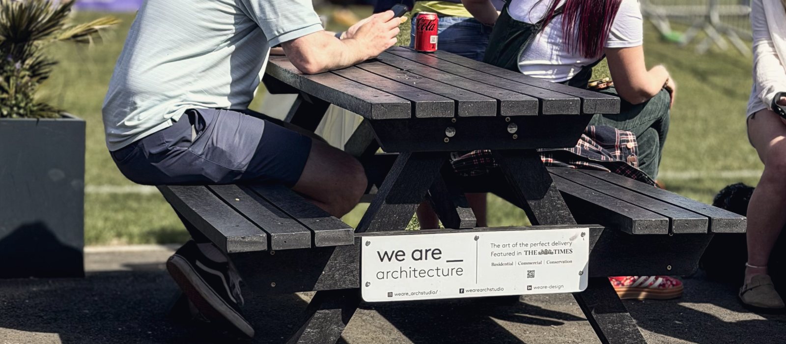 A picnic bench at the cricket club with a we are _ architecture sign fixed to it, people sitting round it in the sun