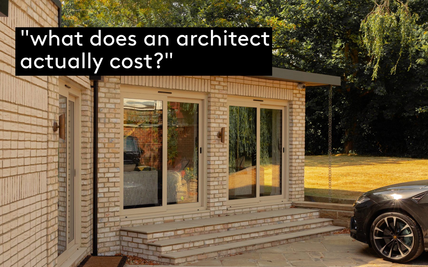 A single-storey brick extension with tall sliding doors opening onto a stone terrace and lawn, seen in low afternoon sun