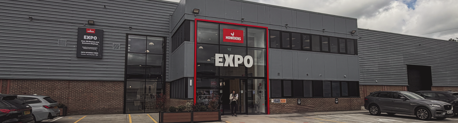 Howdens Expo in Watford, the UK’s first standalone Expo facility — grey profile metal cladding over a brick plinth with a red-framed glazed entrance beacon, brand identity by we are _
