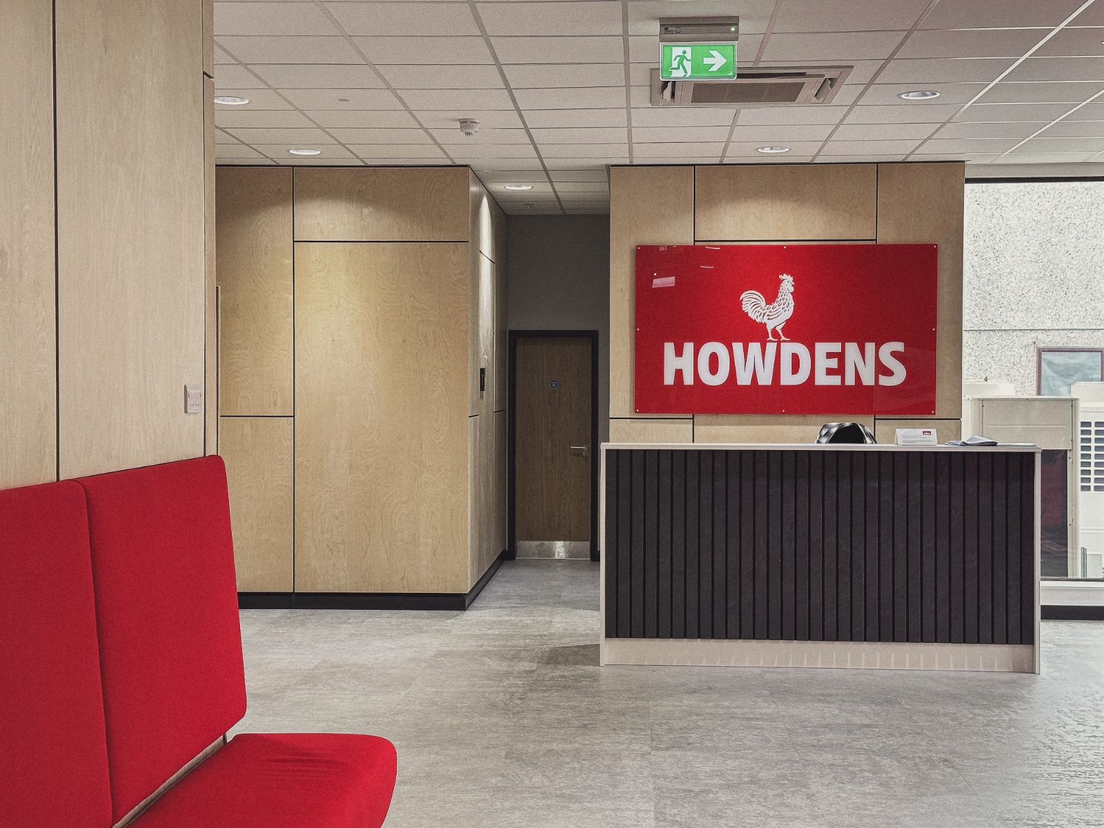 A completed Howdens interior fit-out reception, timber panelling, a slatted desk, a red bench and the red Howdens logo panel, designed and delivered by we are _